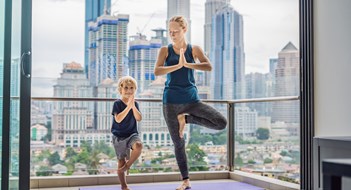 Mom and son are practicing yoga on the balcony in the background of a big city. Sports mom with kid doing morning work-out at home. Mum and child do the exercises together, healthy family lifestyle concept.