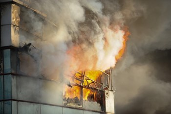 Fire and smoke engulfs a metal and glass office building with the sunset light coming from a side.