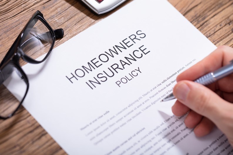 Overhead View Of A Person's Hand Filling Homeowners Insurance Policy Form Over Wooden Desk