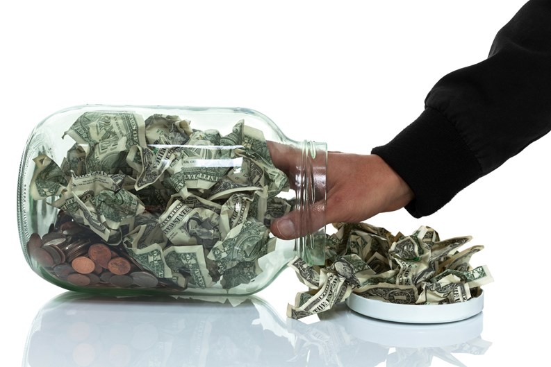 Glass jar full of money tipped over on its side spilling money with hand reaching in