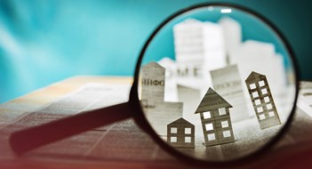 Magnifying glass in front of an open newspaper with paper houses. Concept of rent, search, purchase real estate.