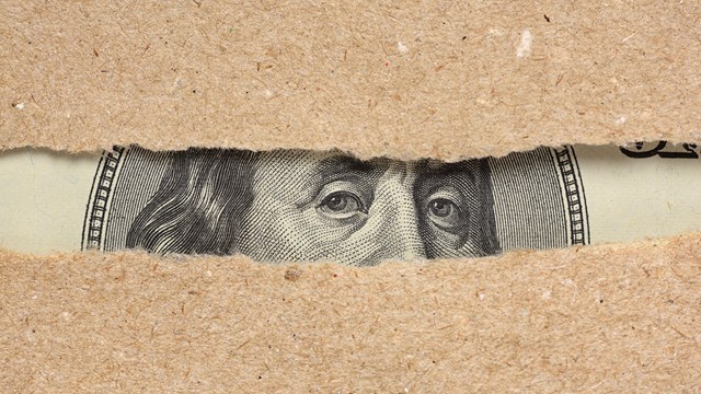 One hundred dollar bill Benjamin Franklin portrait looks behind brown craft ripped paper