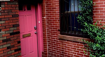 Converting Maisonettes The entrance to a Hoboken, New Jersey basement apartment features a pink door flanked by red and brown brick walls and green vines.