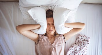 What's That Racket!? Young woman trying to sleep but disturbed by noisy neighbors and covering ears with pillows. I can's sleep with all that noise! Sleep problems. Angry woman disturbed with a noise trying to sleep