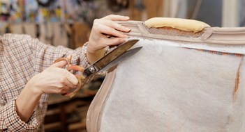 Updating Your Lobby Furnishings Close-up of hands cutting upholstery fabric with large scissors while restoring vintage furniture in a workshop.