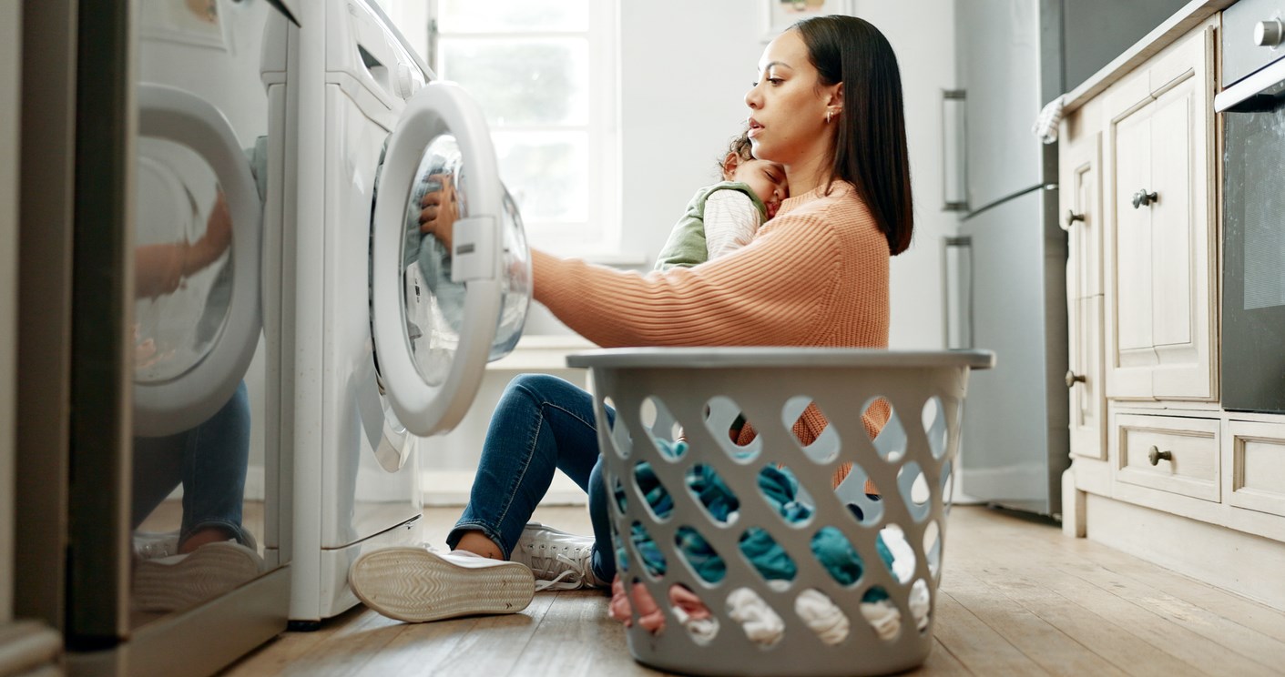 Woman, baby and washing machine on floor, home and sleeping with care, clothes and multitasking. Mother, infant child and basket for laundry, hygiene and cleaning routine in morning at family house