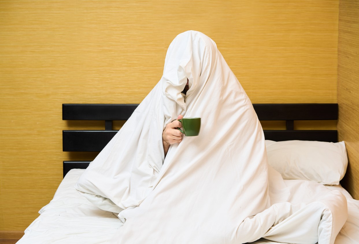 Man wrapping in blanket sitting with cup of coffee in bed. Good morning concept